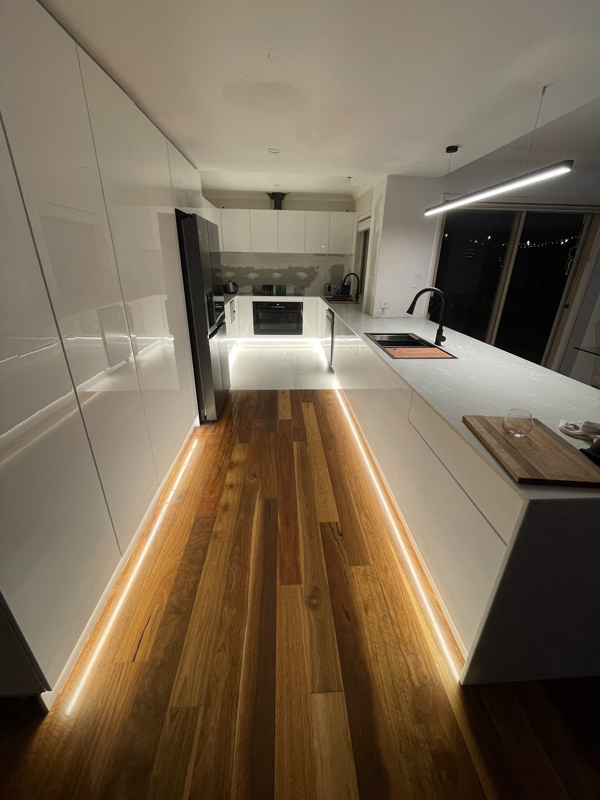Long white kitchen with LED strip lighting glowing along the kickboards and a linear pendant light